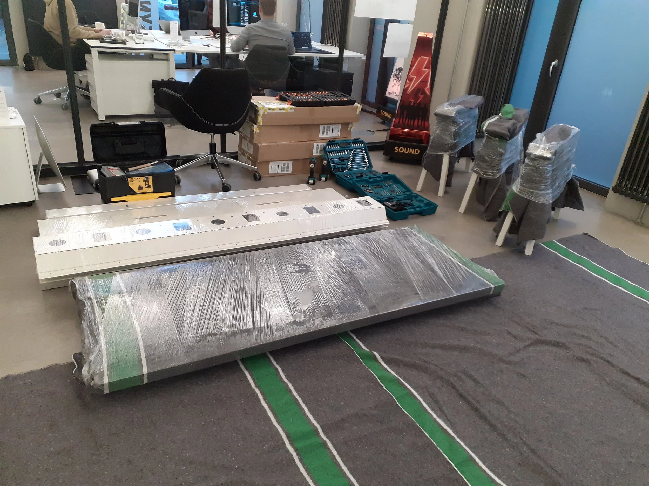 Innenraum eines Büros mit Vorbereitungen für die Installation eines großen LED-Bildschirms, mit Werkzeugen, Verpackungsmaterial, Stühlen und Arbeitstischen.