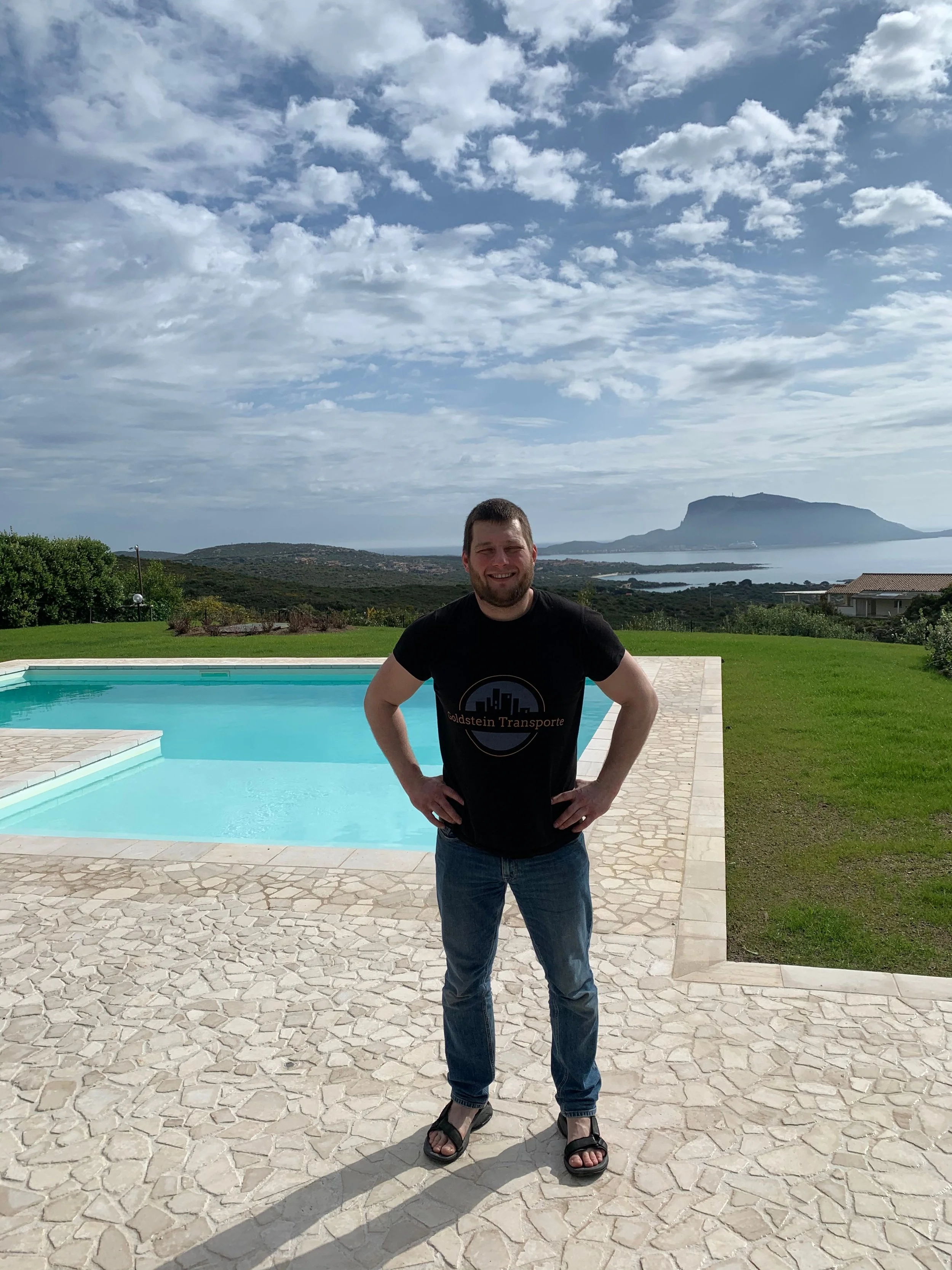 Mann steht vor einem swimming pool mit Blick auf das Meer und eine Insel im Hintergrund, während der Himmel teilweise bewölkt ist.