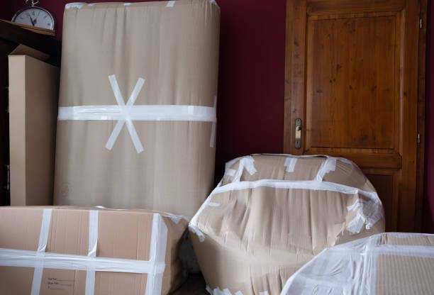 Vier große Gegenstände, vermutlich Möbelstücke, sind mit braunem Packpapier und weißen Klebeband in einem Raum verpackt, neben einer Holztür.