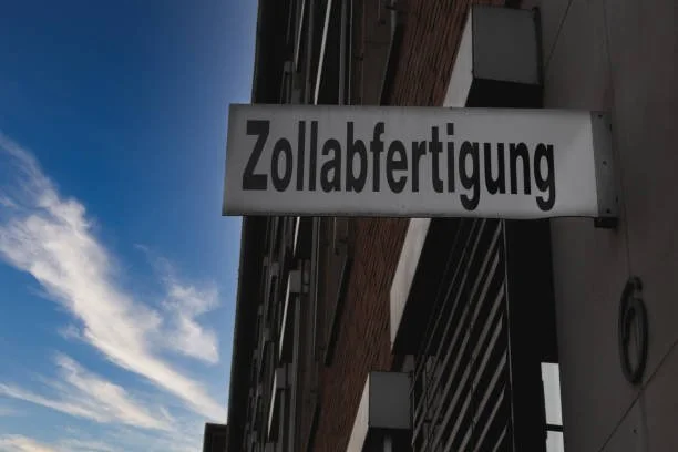 Schild mit der Aufschrift 'Zollabfertigung' an einem Gebäude, Himmel mit Wolken im Hintergrund.