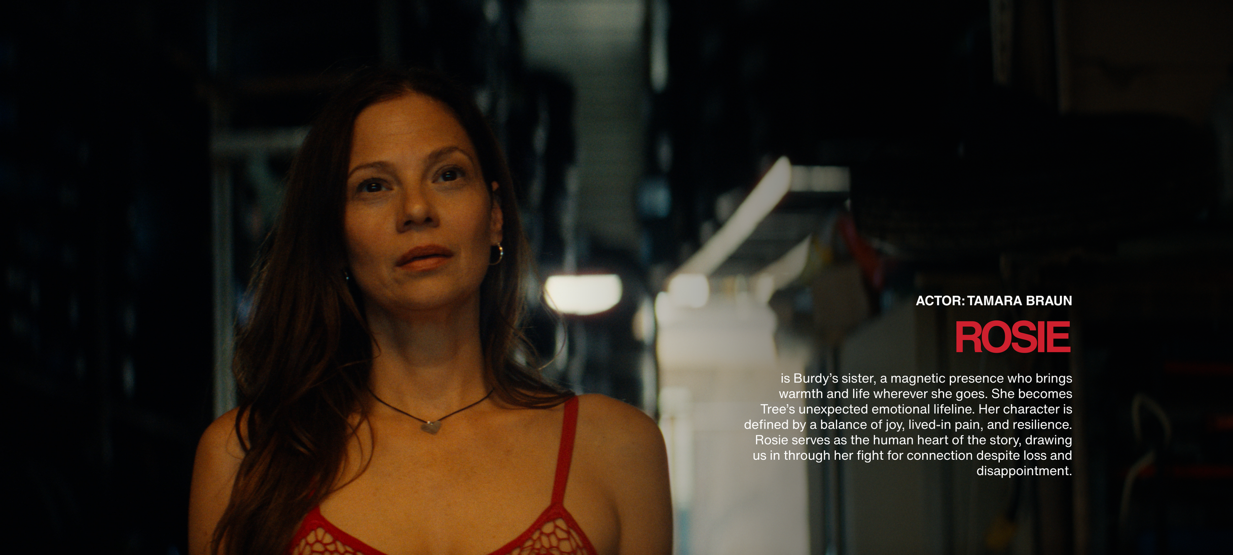 A woman with long brown hair wearing a red strappy top and a heart-shaped necklace, standing in a dimly lit industrial setting.