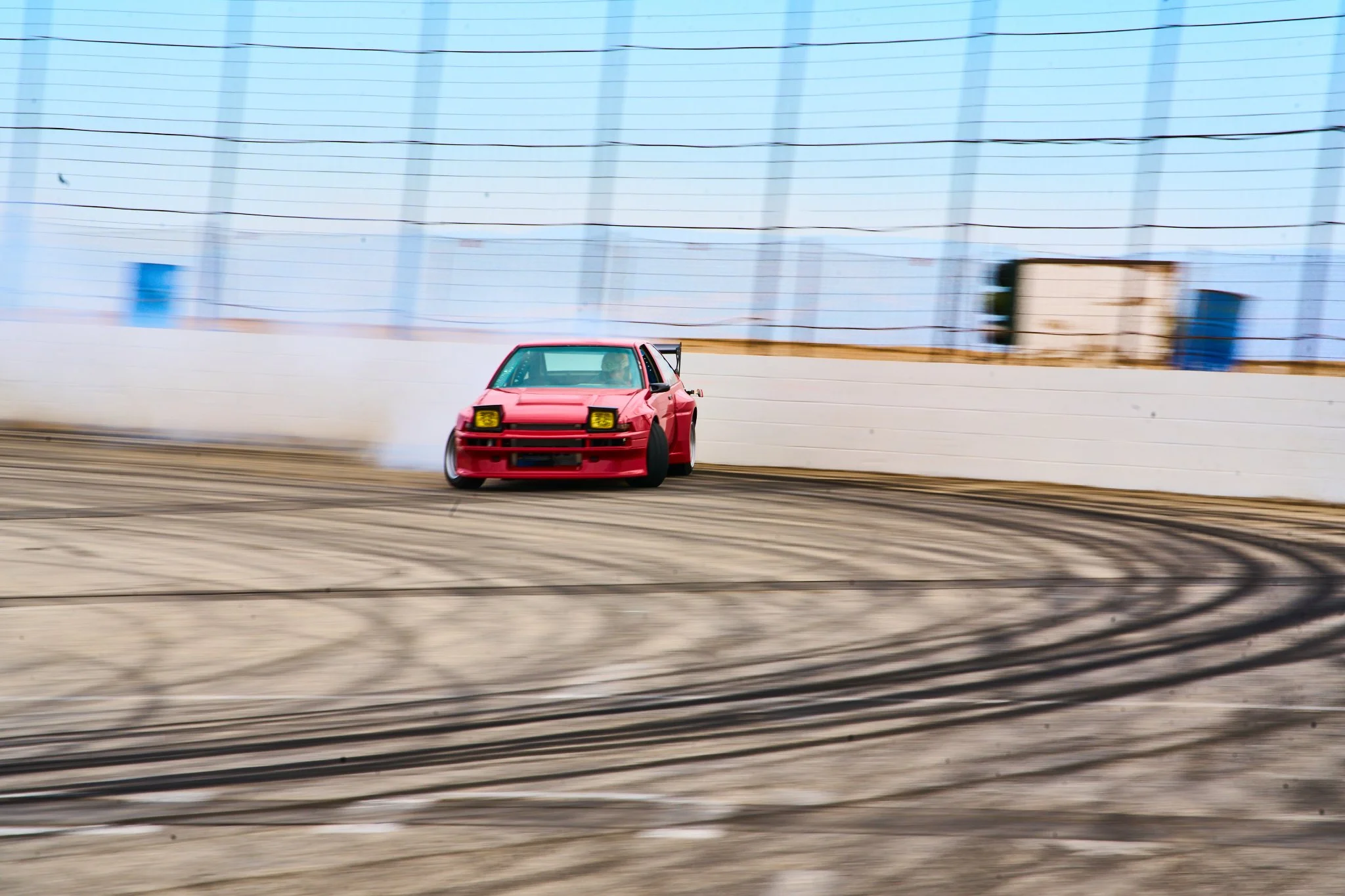 A red race car drifting on a race track with tire smoke and skid marks, surrounded by a tall blue fence.