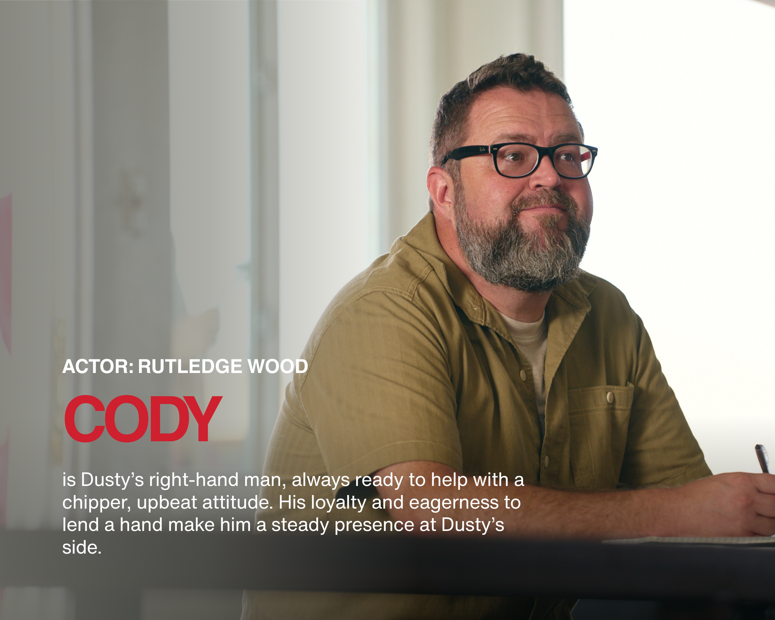 A man with glasses and a beard, wearing a brown shirt, sitting at a desk in a well-lit room with a window behind him.