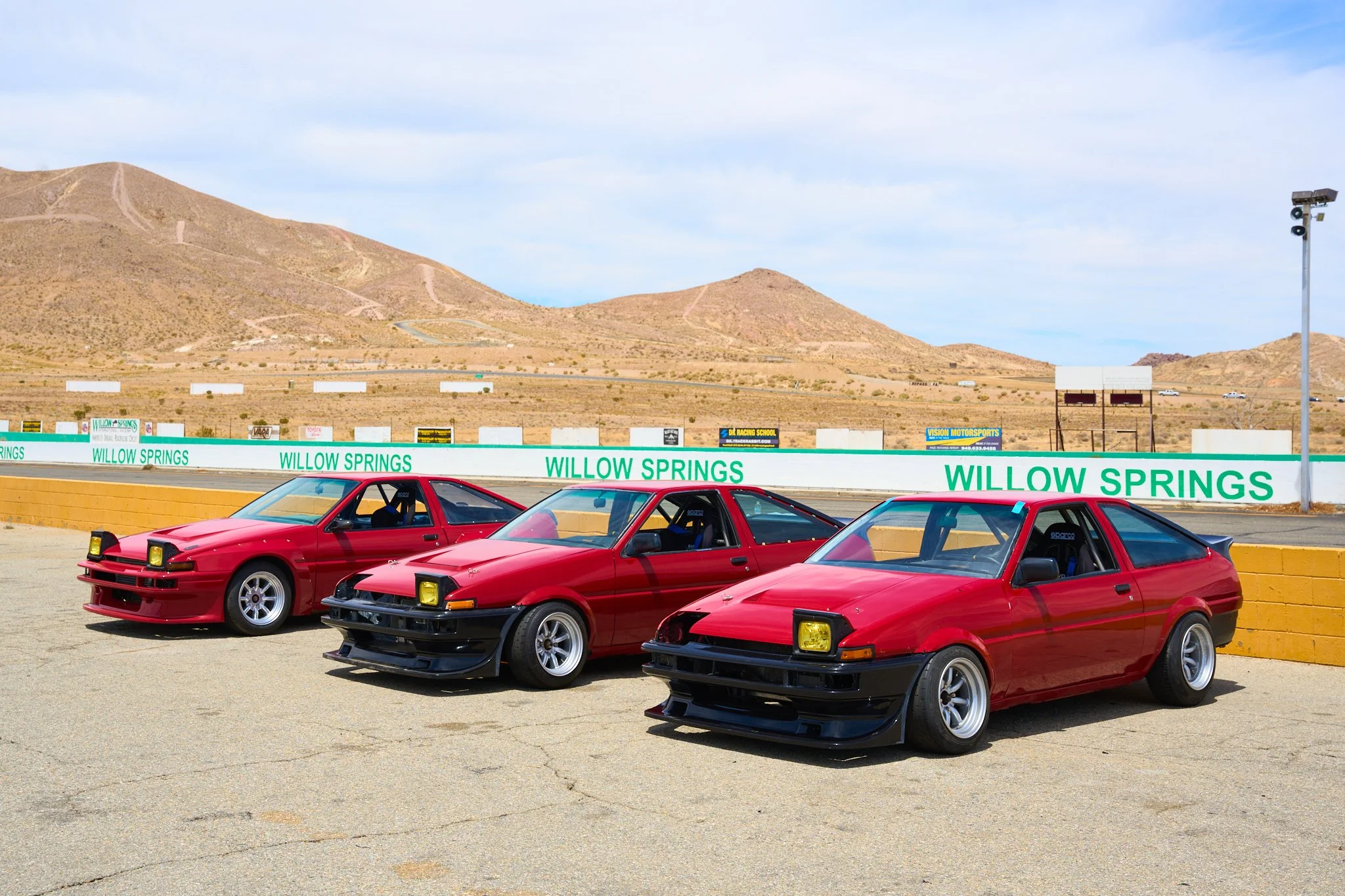 Three red vintage sports cars are parked side by side on a race track with a desert mountain landscape in the background, and a large sign reading 'Willow Springs' along the track's wall.