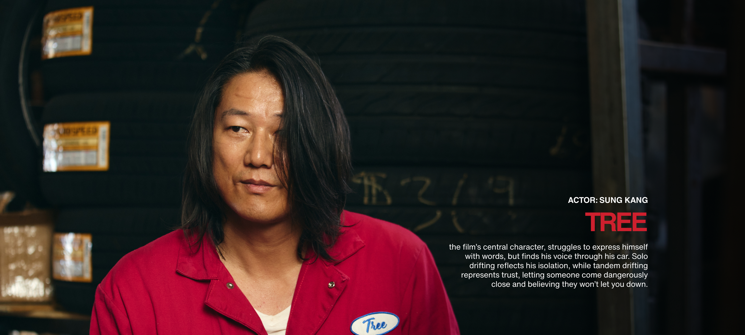 Close-up of a man with long black hair, wearing a red work uniform with a "Tree" name tag, in a tire shop with stacks of tires in the background. Text overlay indicating actor Sung Kang and the character "Tree."