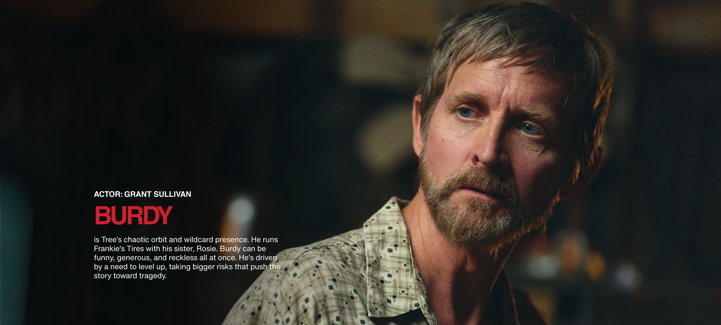 Close-up of actor Grant Sullivan with a serious expression, wearing a plaid shirt, in a dimly lit setting.
