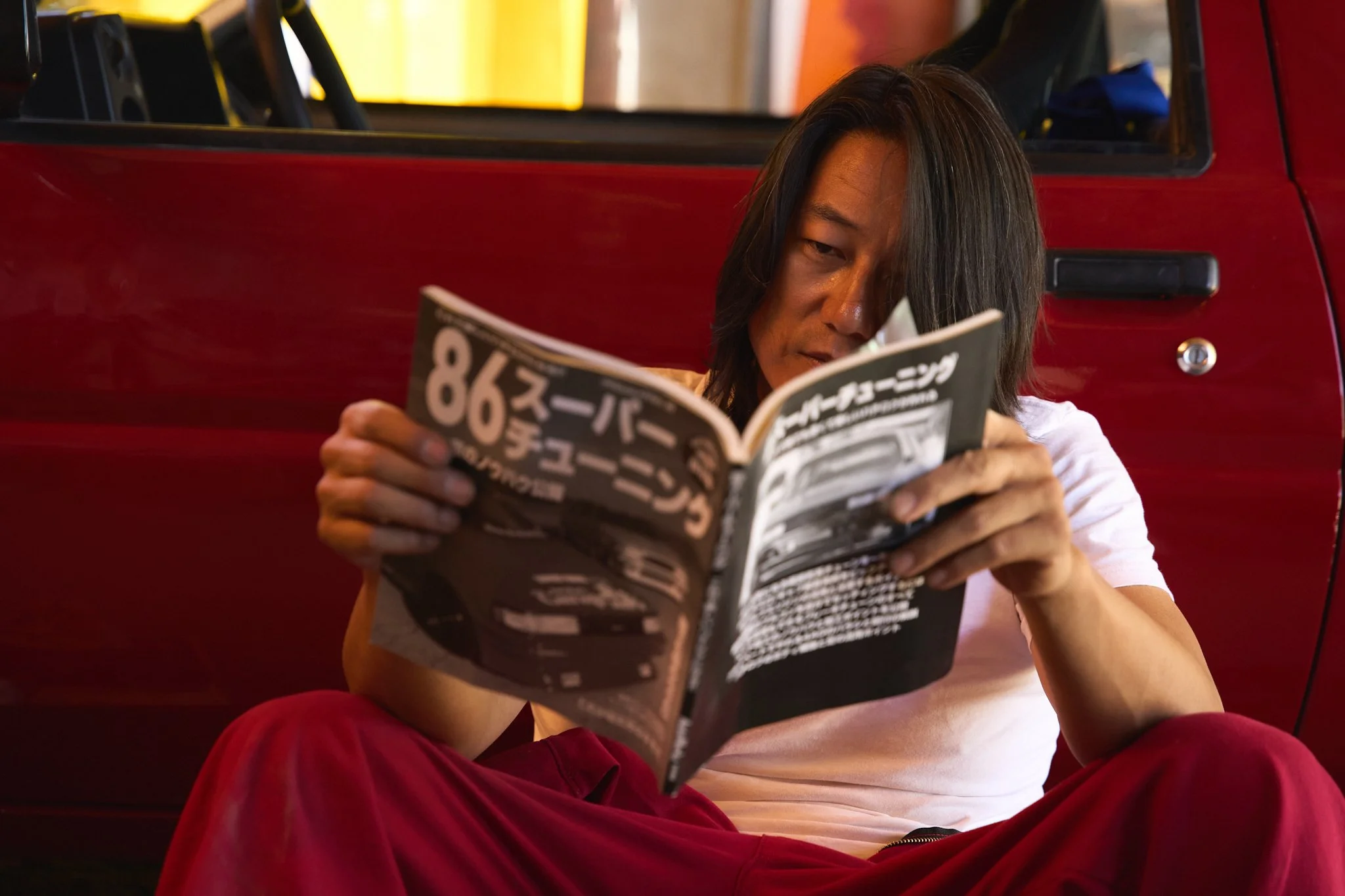 A person with long dark hair sitting on the ground reading a magazine in front of a red vehicle.