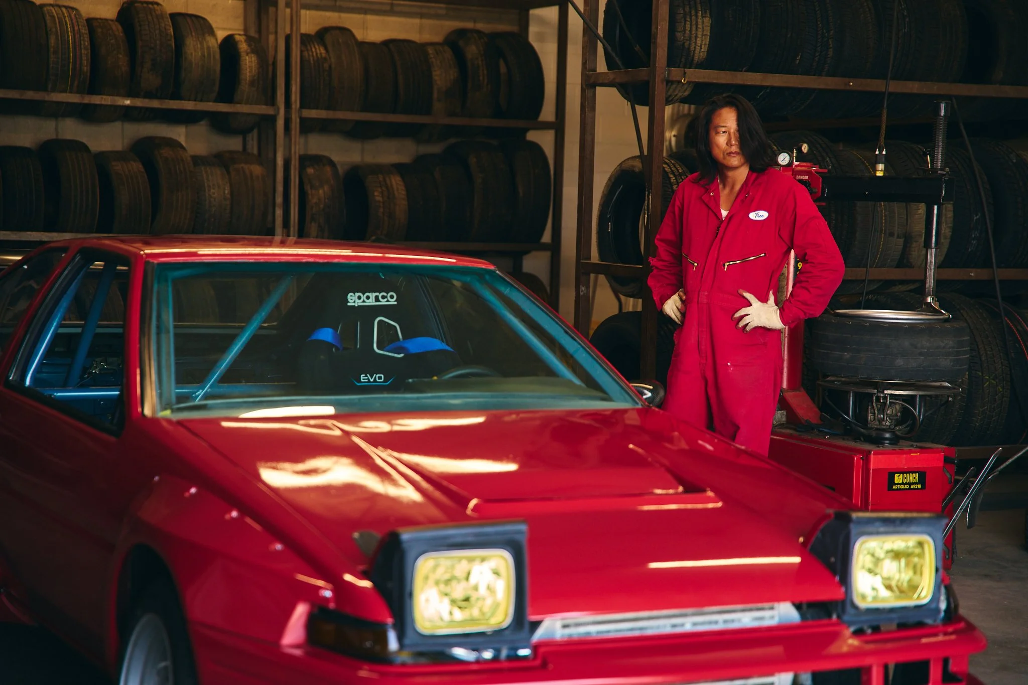 A man in a red jumpsuit standing next to a red race car inside a garage, with tires stacked on shelves behind him.