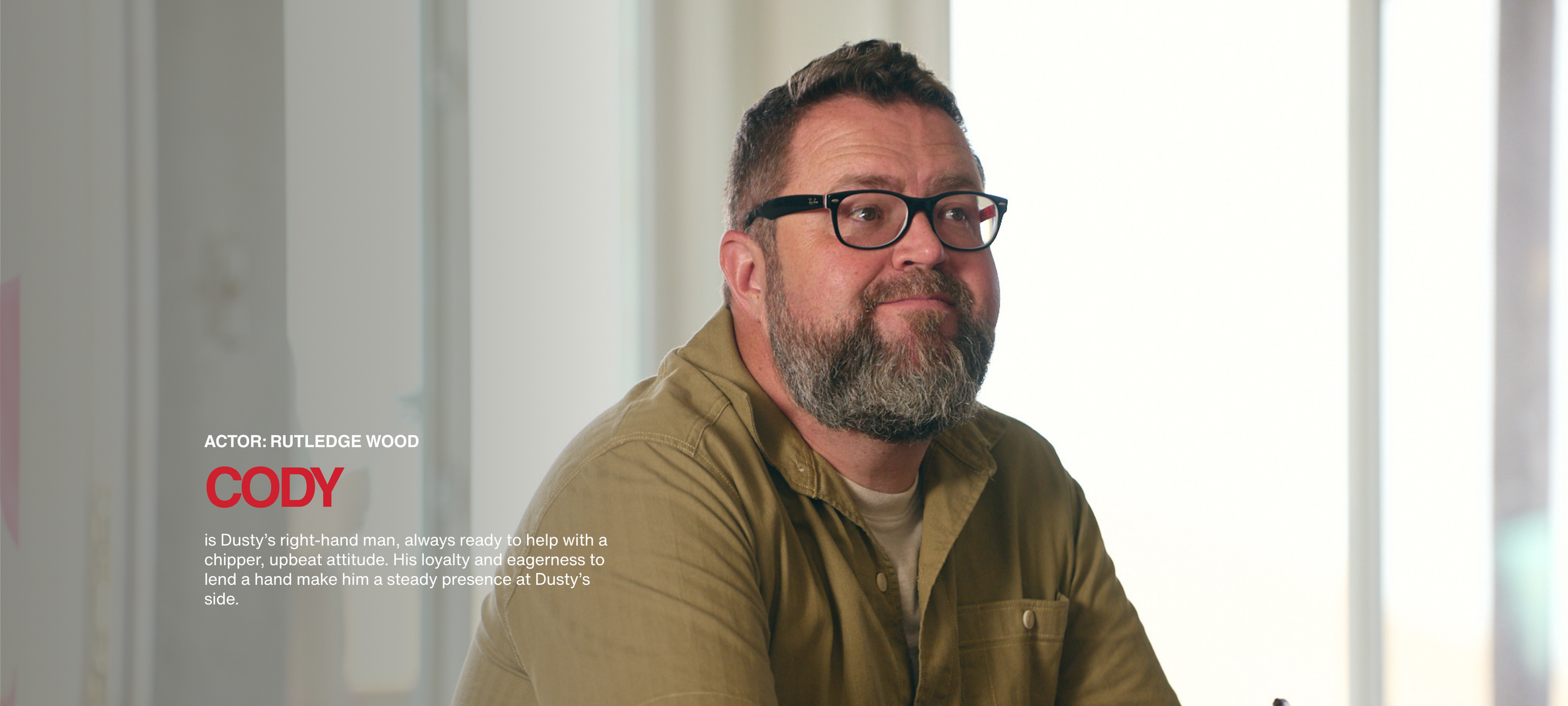 A middle-aged man with glasses and a beard, wearing a tan shirt, sitting indoors near a window with bright light.