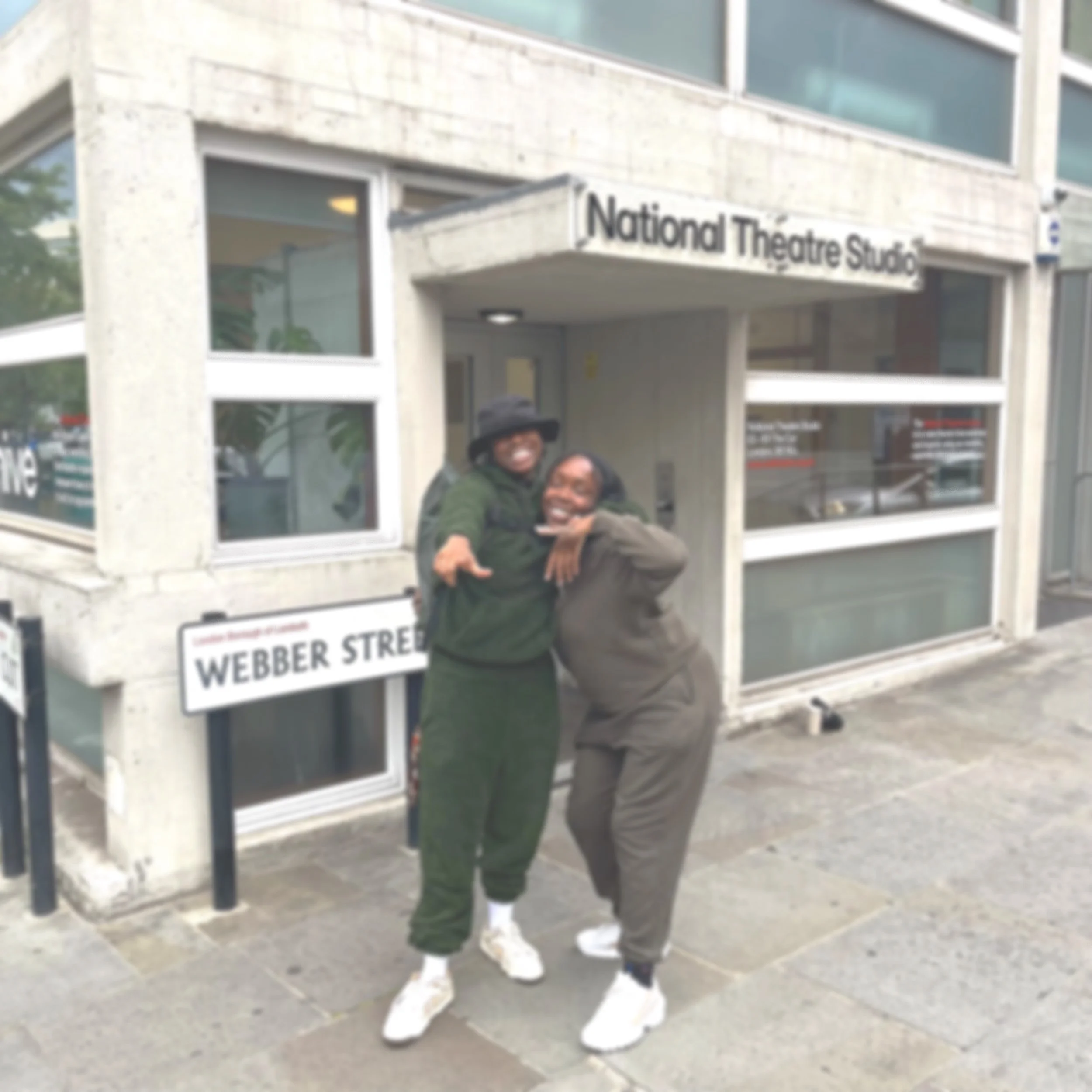 Two people smiling and posing joyfully outside a building labeled as National Theatre Studio, on Webber Street, with one wearing a dark hat and glasses, and the other in a brown jacket and pants.