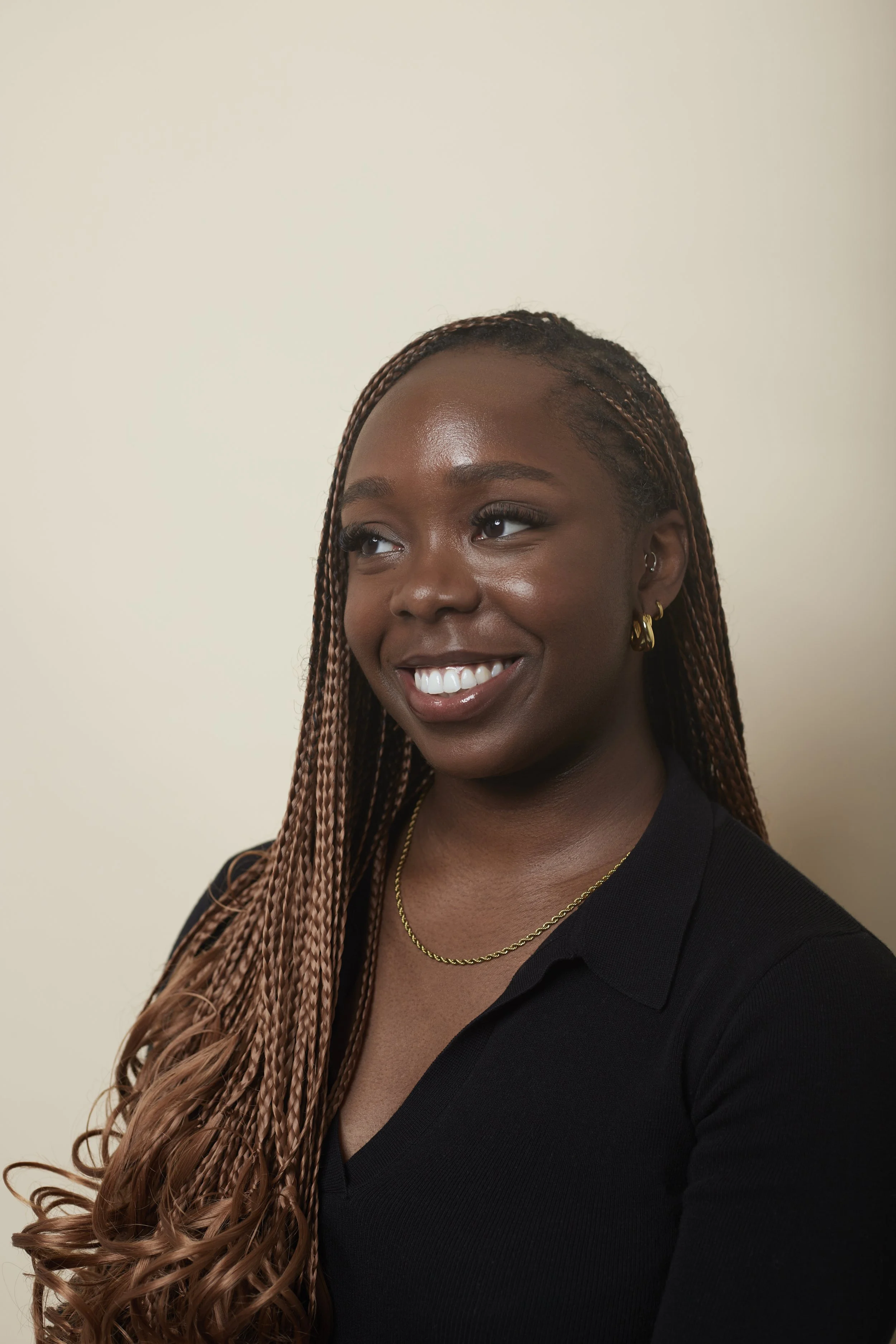 A young woman with long, twisted hair smiling and wearing a black top, gold jewelry, and earrings, against a plain light-colored background.