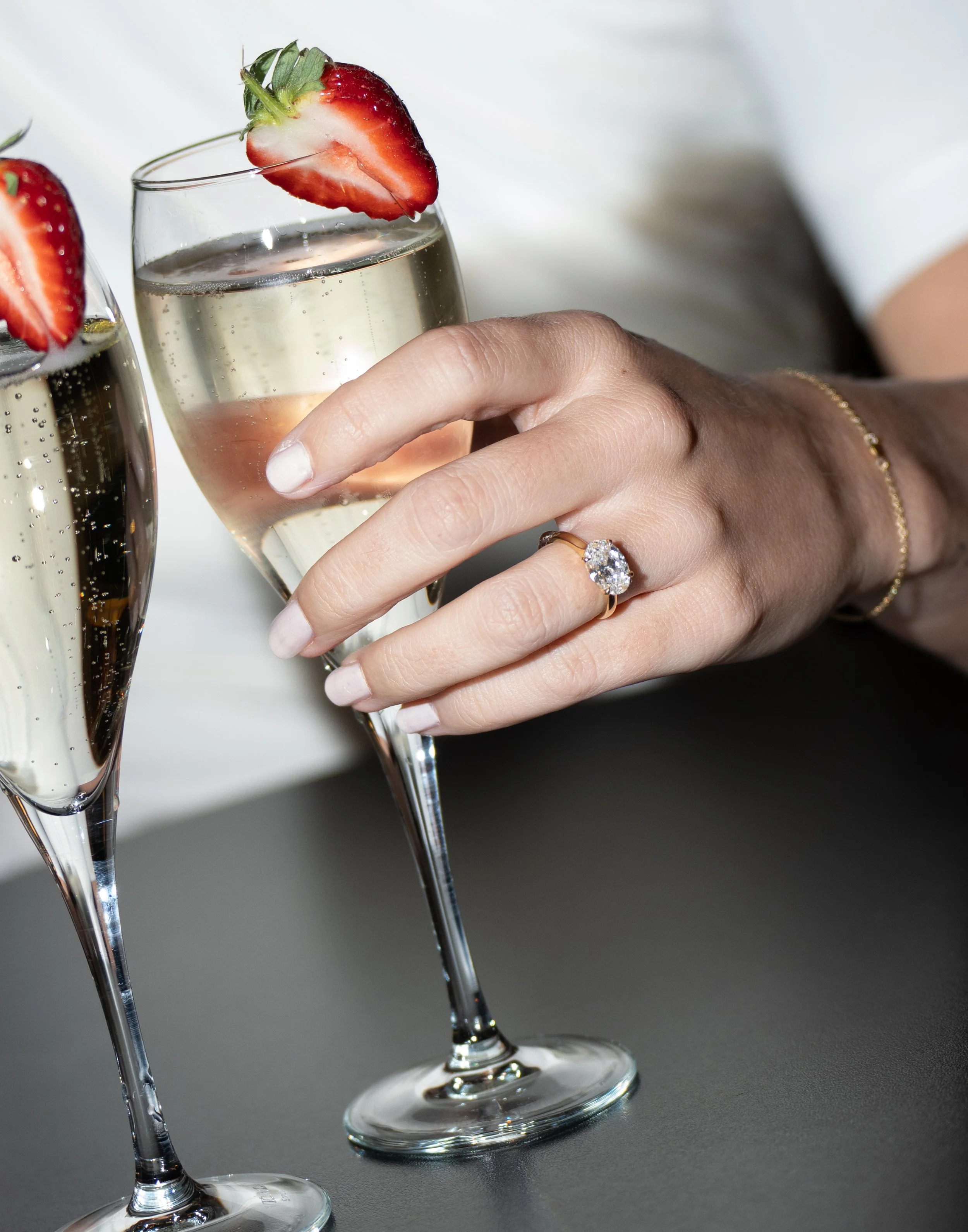 Two champagne glasses filled with a clear sparkling beverage, garnished with halved strawberries on the rim, resting on a dark surface with a blurred background.