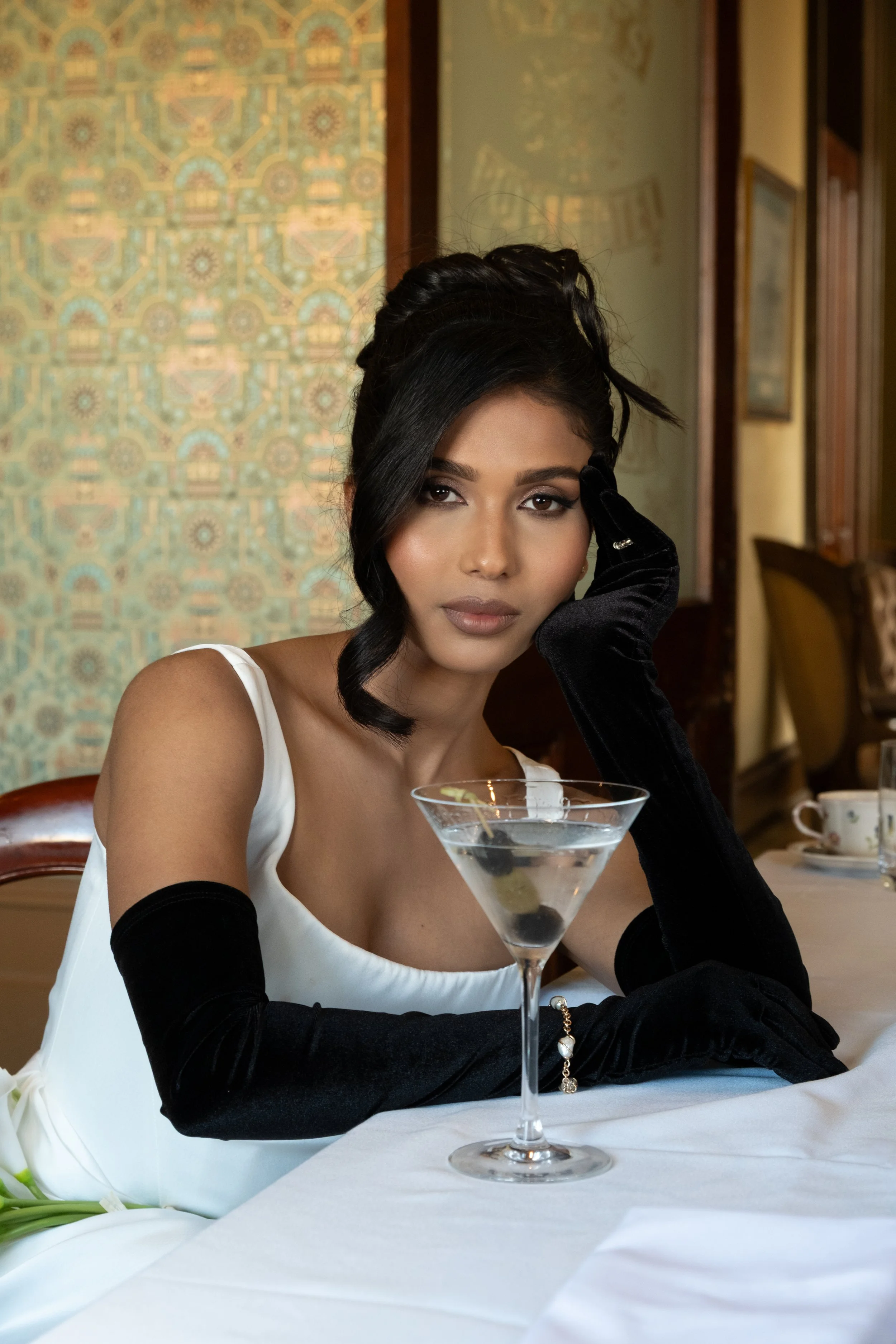 A woman with dark hair styled in an elegant updo, wearing a white sleeveless dress and black gloves, sitting at a table in a fancy restaurant. She is resting her head on her hand while looking at the camera, with a martini glass with olives in front of her. The background shows a patterned wall and framed pictures.