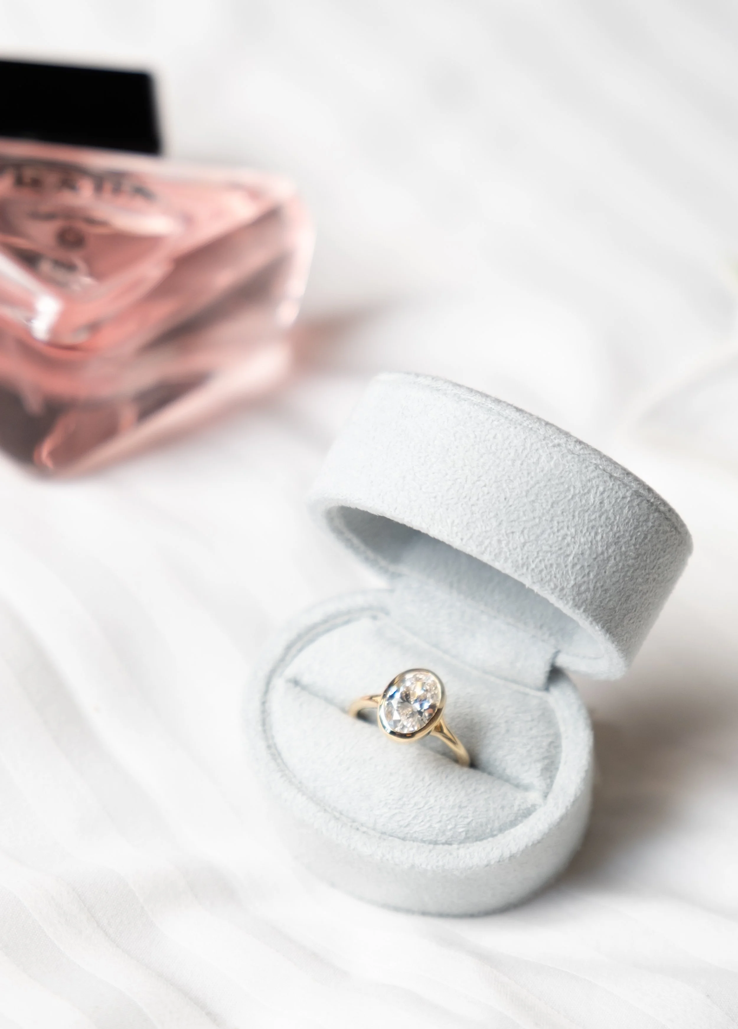 Close-up of a ring with a large clear gemstone in a white padded ring box, with a pink jewelry box partially visible in the background.