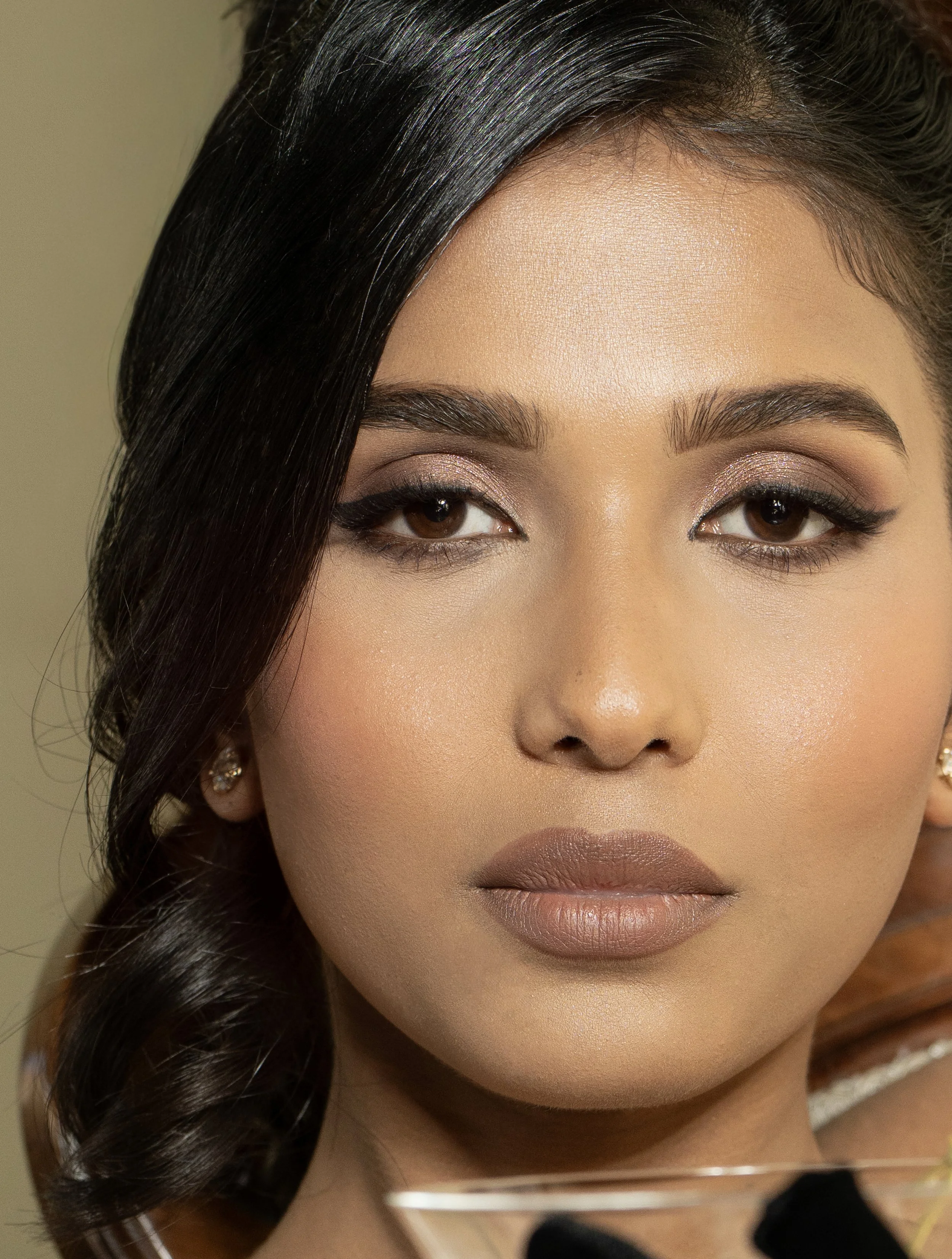 Close-up of a woman's face showing her full makeup, including eye makeup with eyeshadow and eyeliner, well-groomed eyebrows, and matte lipstick, with dark, shiny hair styled to one side.