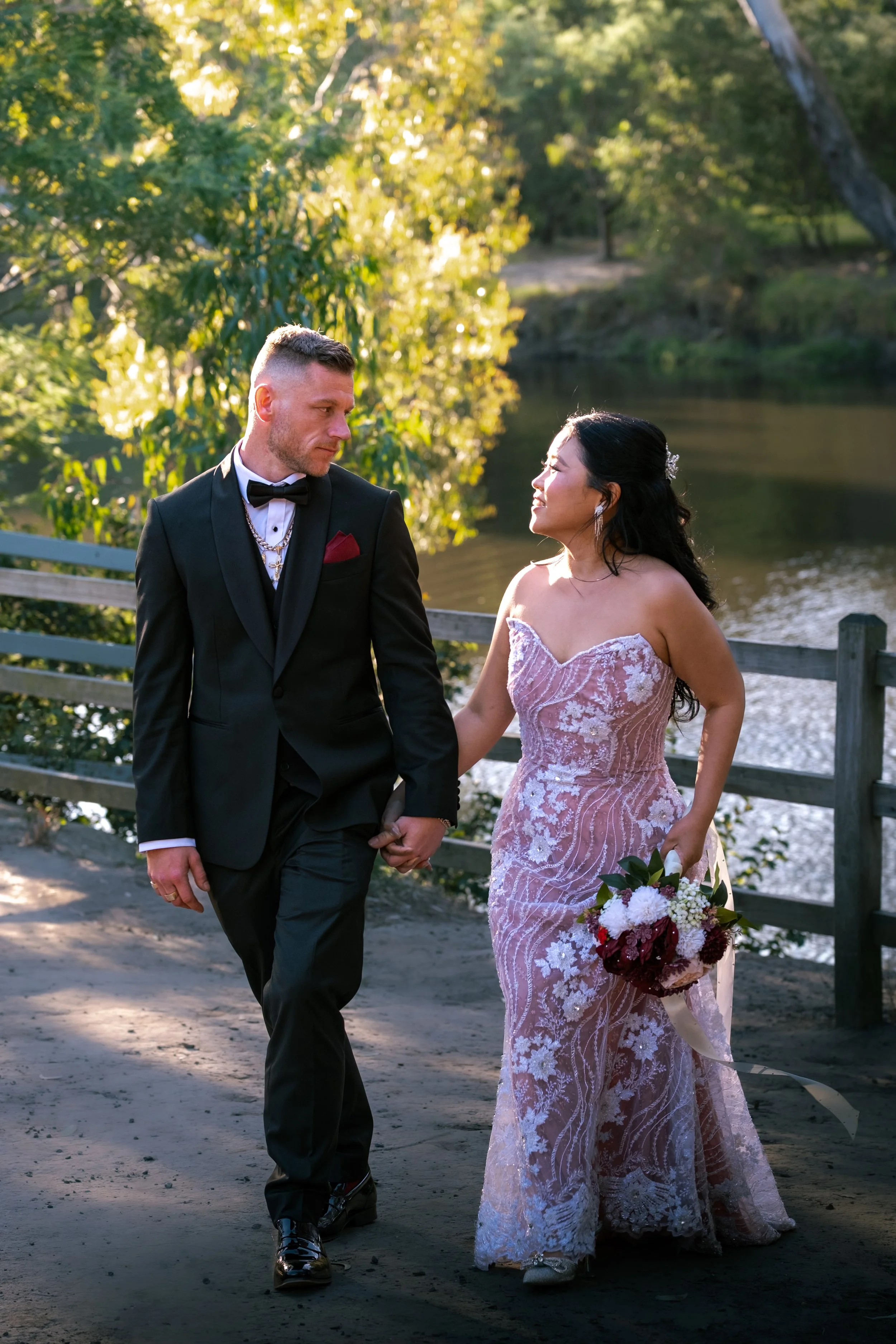 A couple in wedding attire walking hand in hand near a river surrounded by trees during sunset.