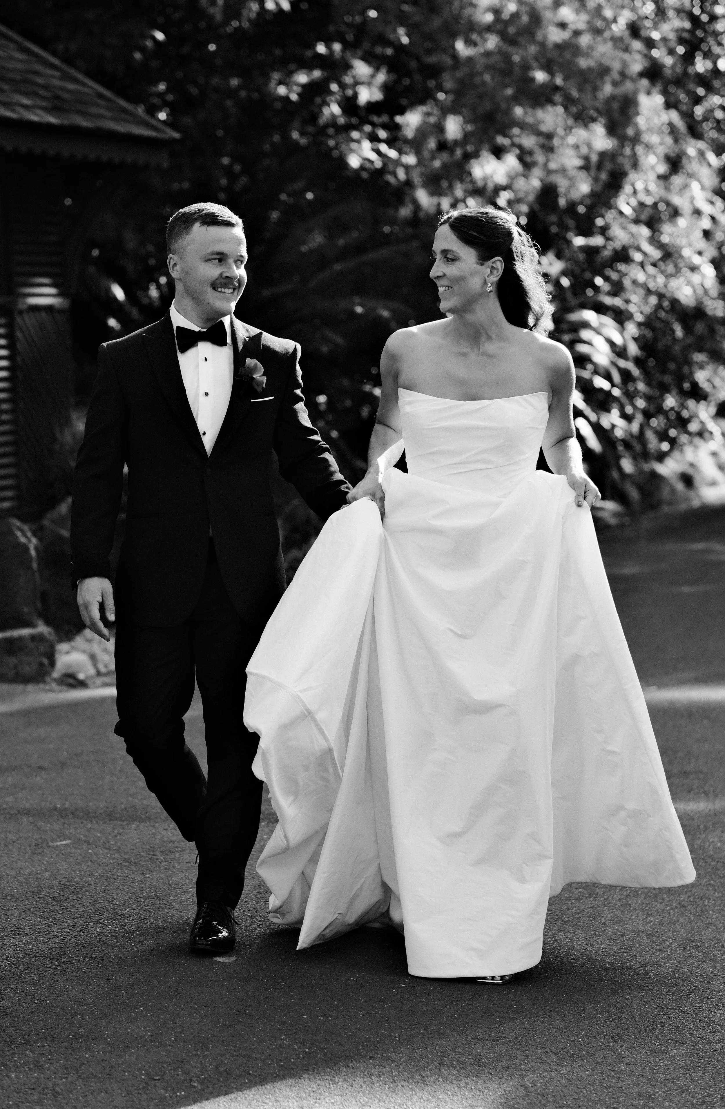 A bride and groom are walking outside, holding hands and smiling at each other during a wedding. The bride is in a strapless white wedding gown, and the groom is in a black tuxedo with a bowtie.