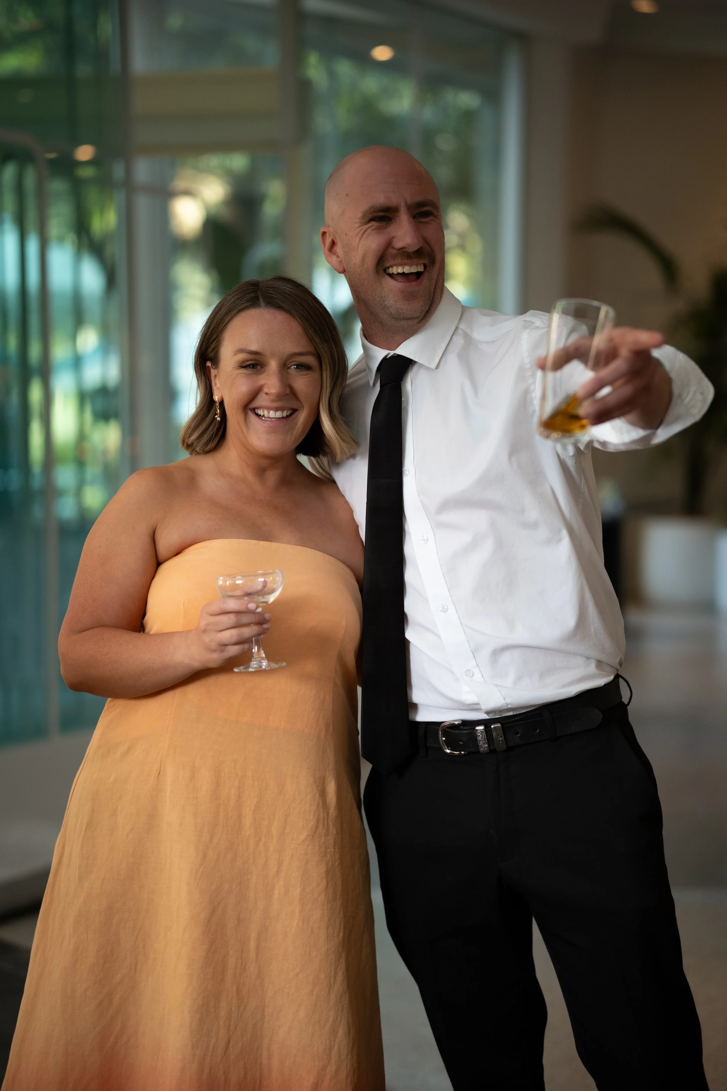 Two people are smiling and toasting with drinks in an indoor setting with large windows and greenery outside.