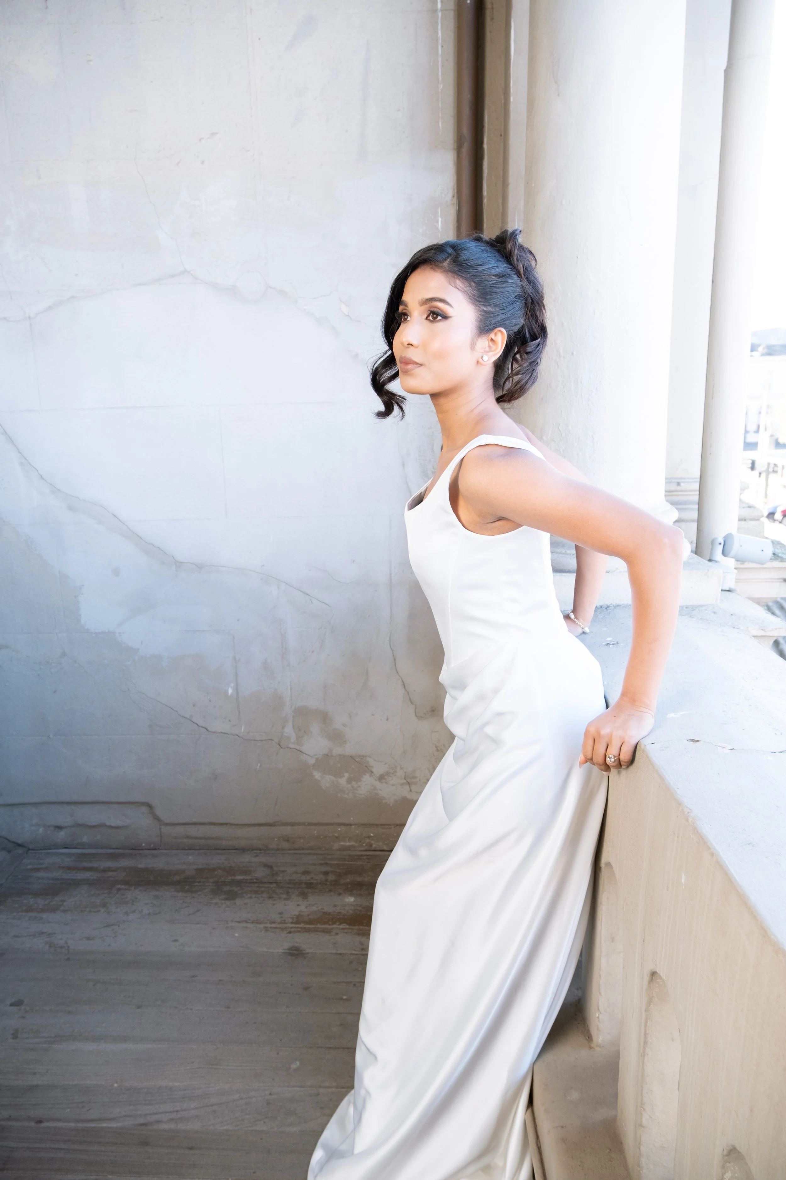A woman in a white dress standing against a textured wall and leaning on a ledge outside a historic building with columns.