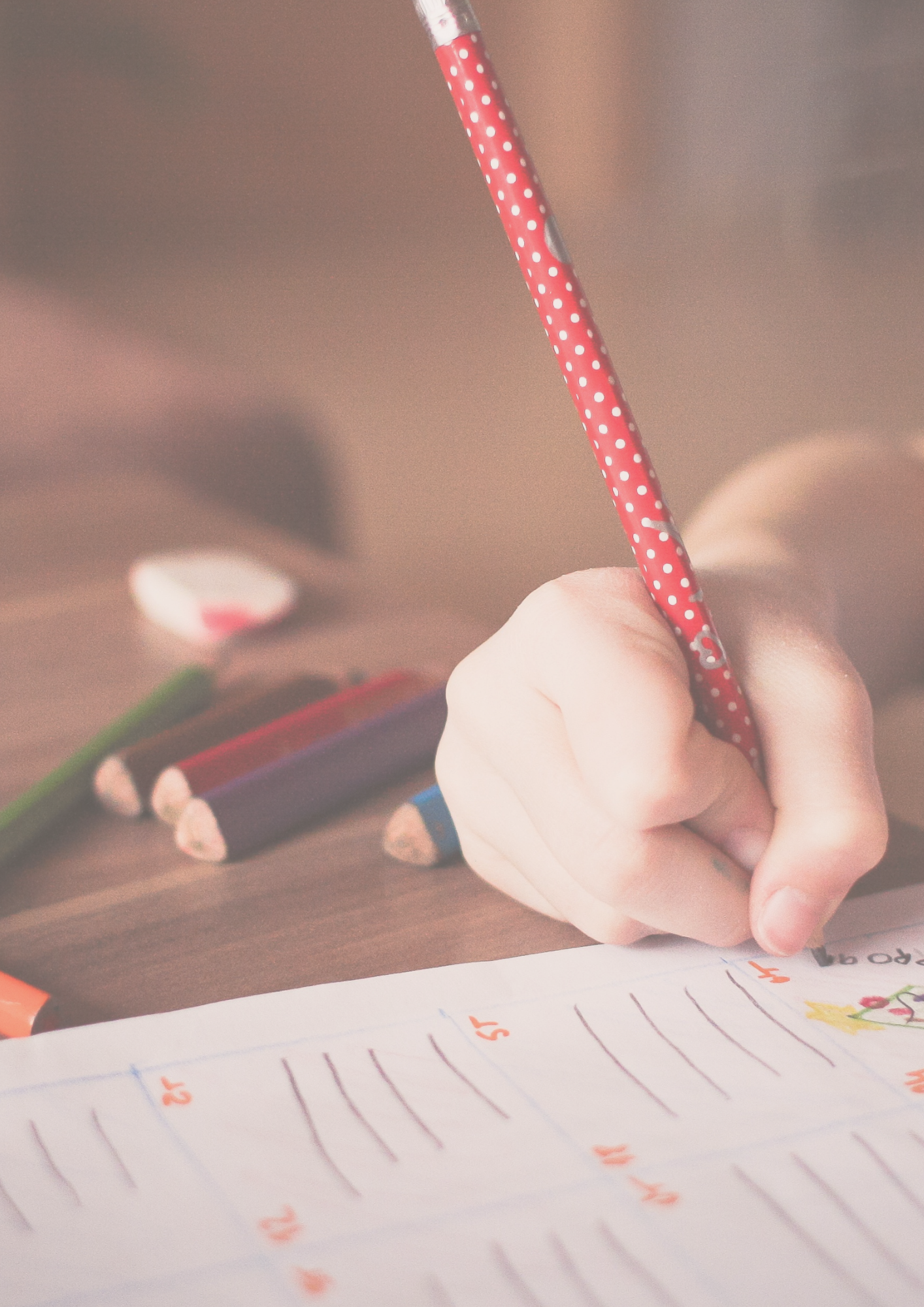 A child's hand holding a red polka-dotted pencil, writing on a worksheet with colorful pencils and an eraser nearby on a wooden table.