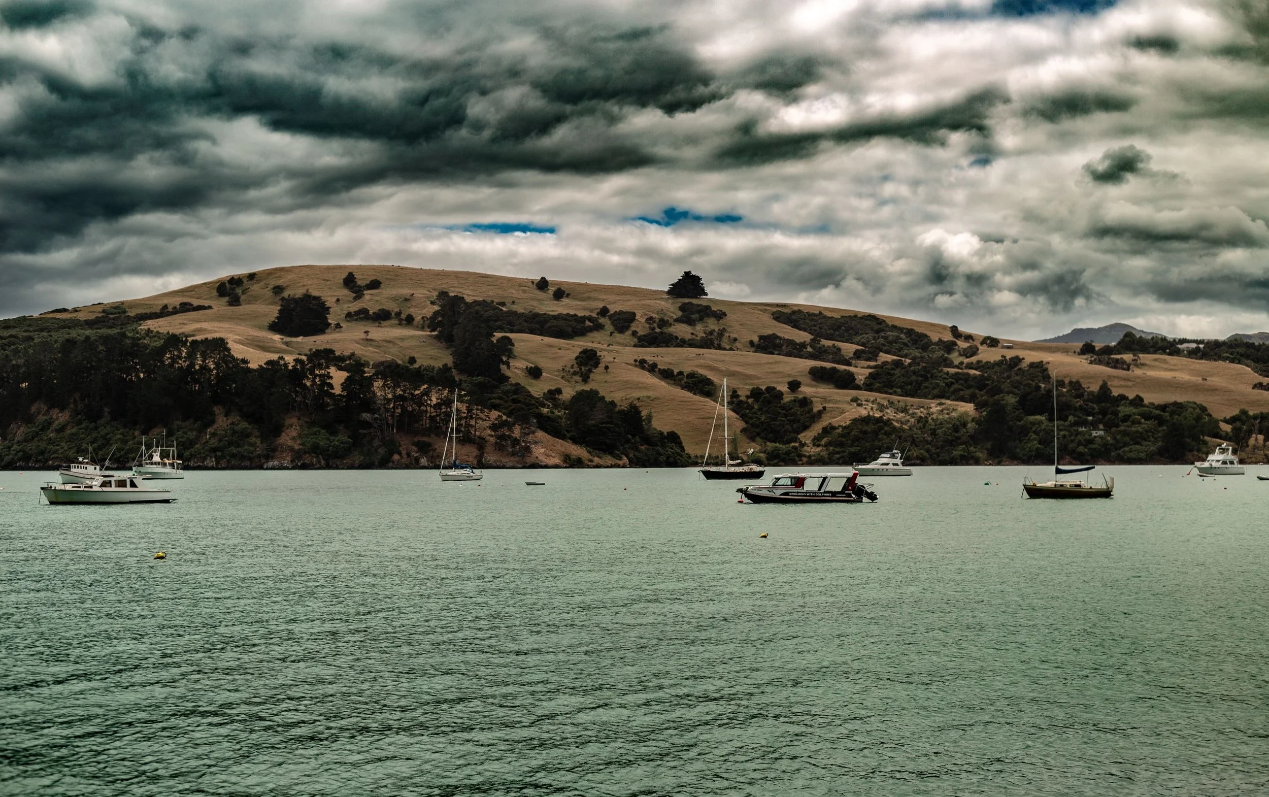 Boats floating on the water of a bay with rolling hills and cloudy sky in the background.
