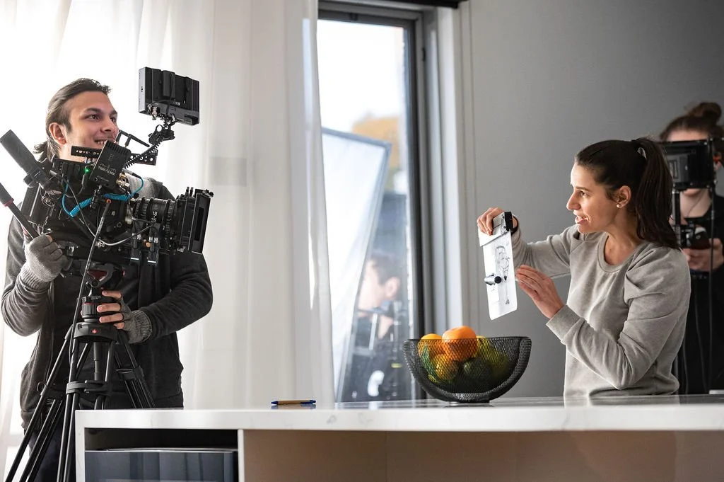 Filming crew on set, with a woman holding a clapboard and a man operating a camera on a tripod in a bright kitchen.