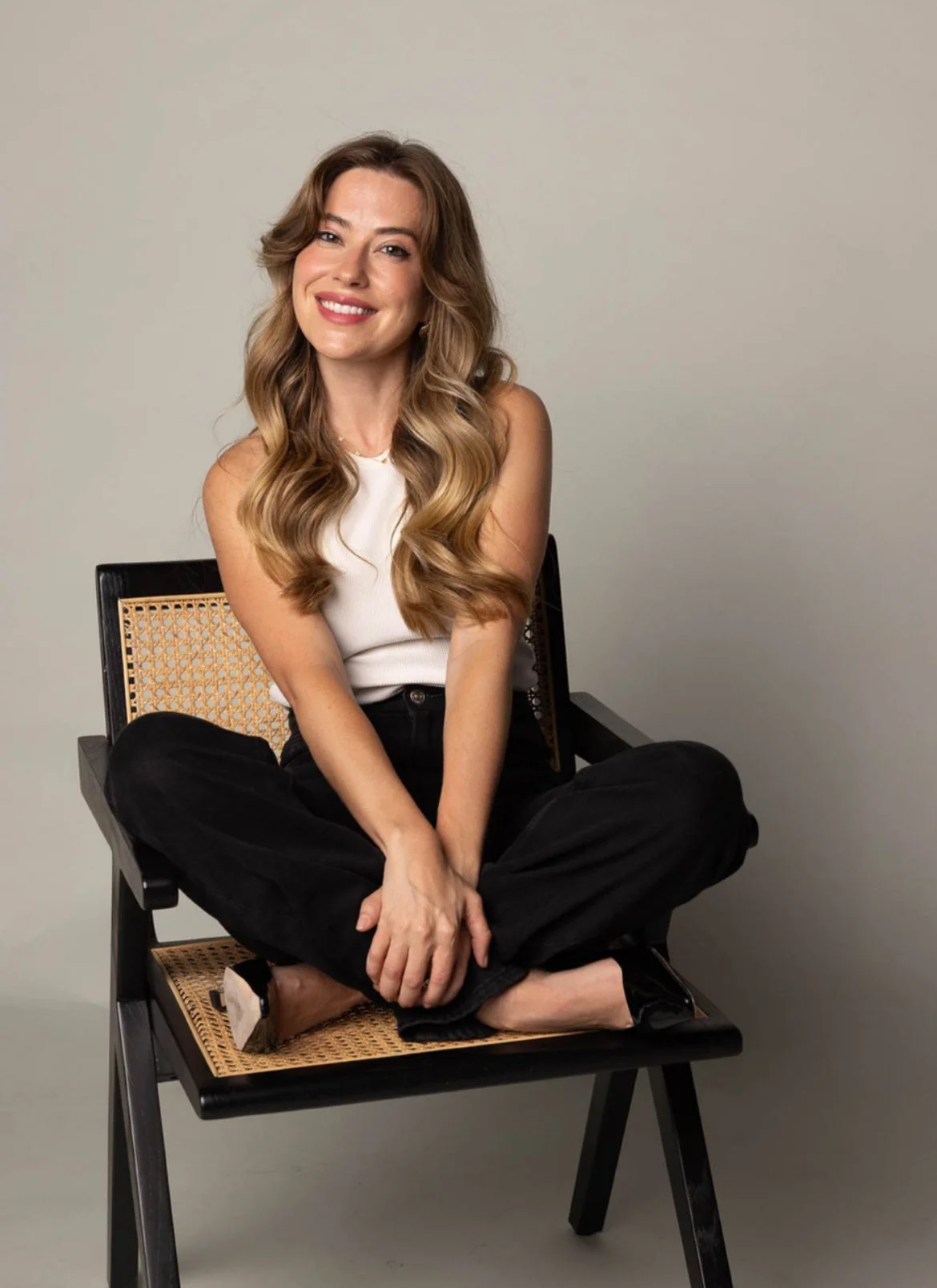 A woman with long wavy brown hair, smiling, sitting cross-legged on a black chair with a woven back and seat, against a plain off-white background.