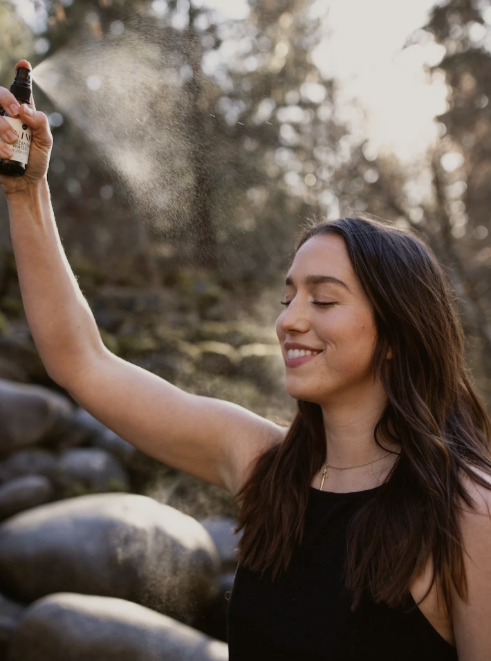 Young woman outdoors smiling as she sprays a mist from a small bottle.