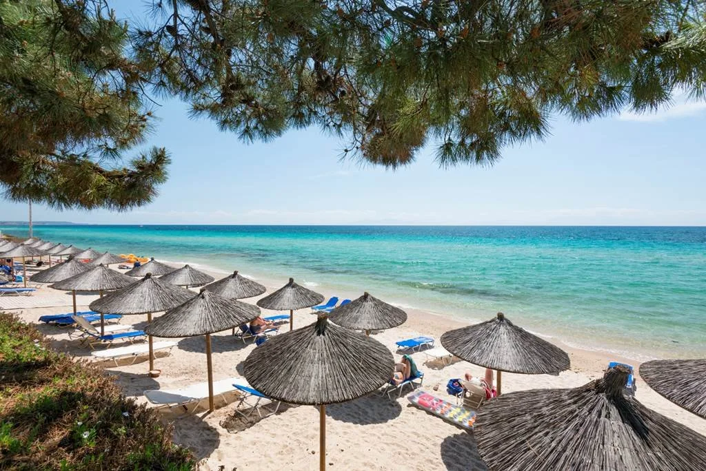 A beach scene with multiple straw umbrellas, lounge chairs, and people relaxing on the white sandy beach under a partially shaded area with a view of calm turquoise ocean and a clear blue sky.