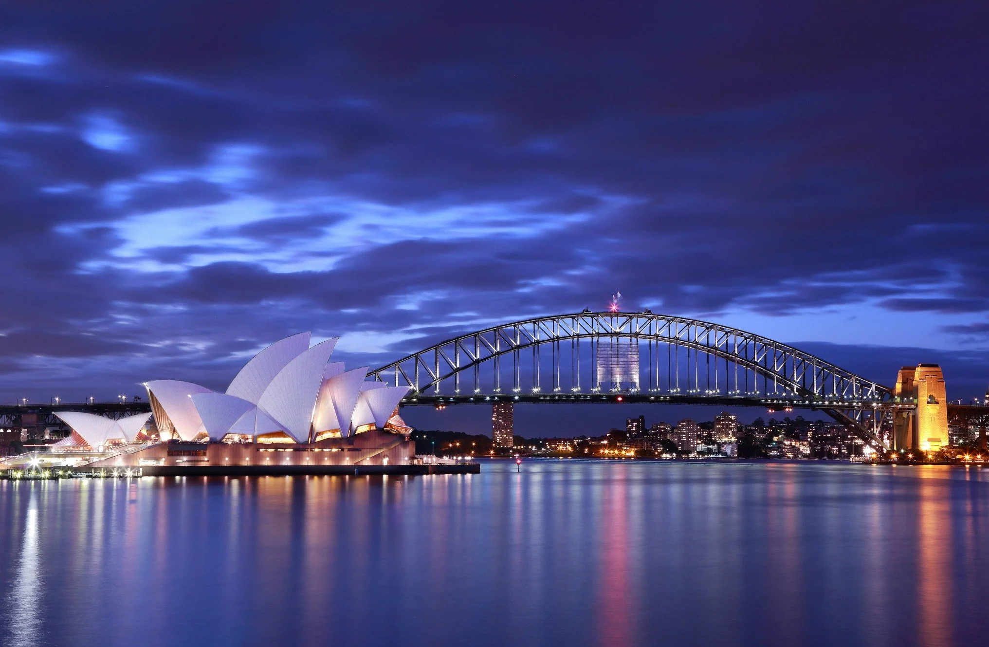 Night view of the Sydney Opera House and Harbour Bridge illuminated, reflecting on the water.