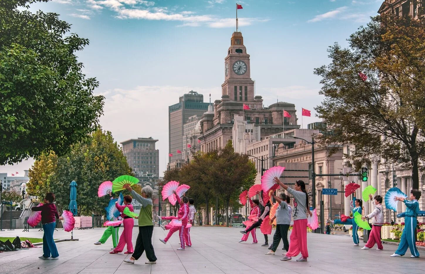 Group of people practicing fan dance in a city square, with historic building featuring a clock tower and pink flags in the background, early evening with clear sky.