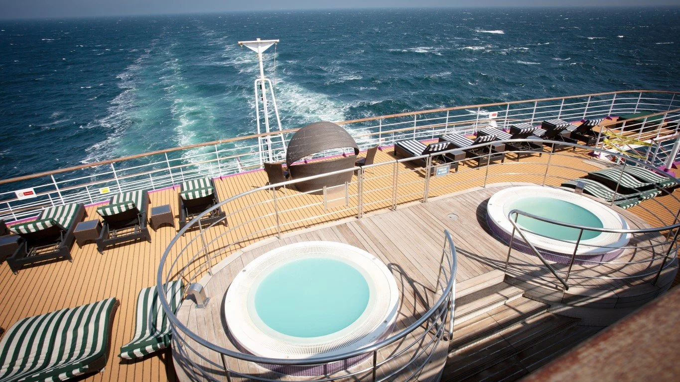 View of a cruise ship deck with two small hot tubs, lounge chairs, and an ocean view with a wake trail behind the ship.