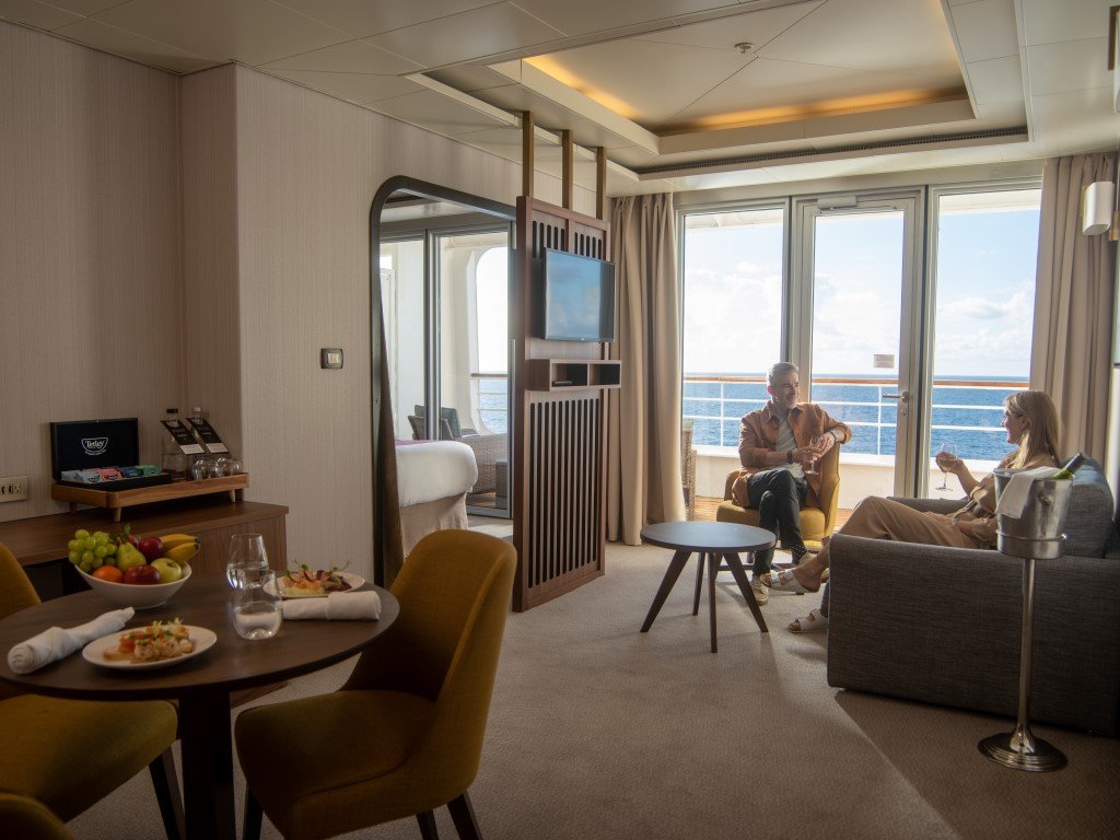 Two people sit and chat on a balcony over the ocean, visible through large closed glass doors, inside a hotel room with a dining area and living space.