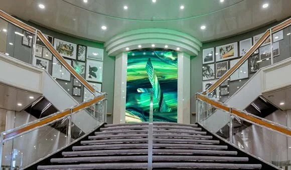 Interior of a modern building with a staircase leading up to a large digital display at the top, surrounded by framed pictures and artwork.
