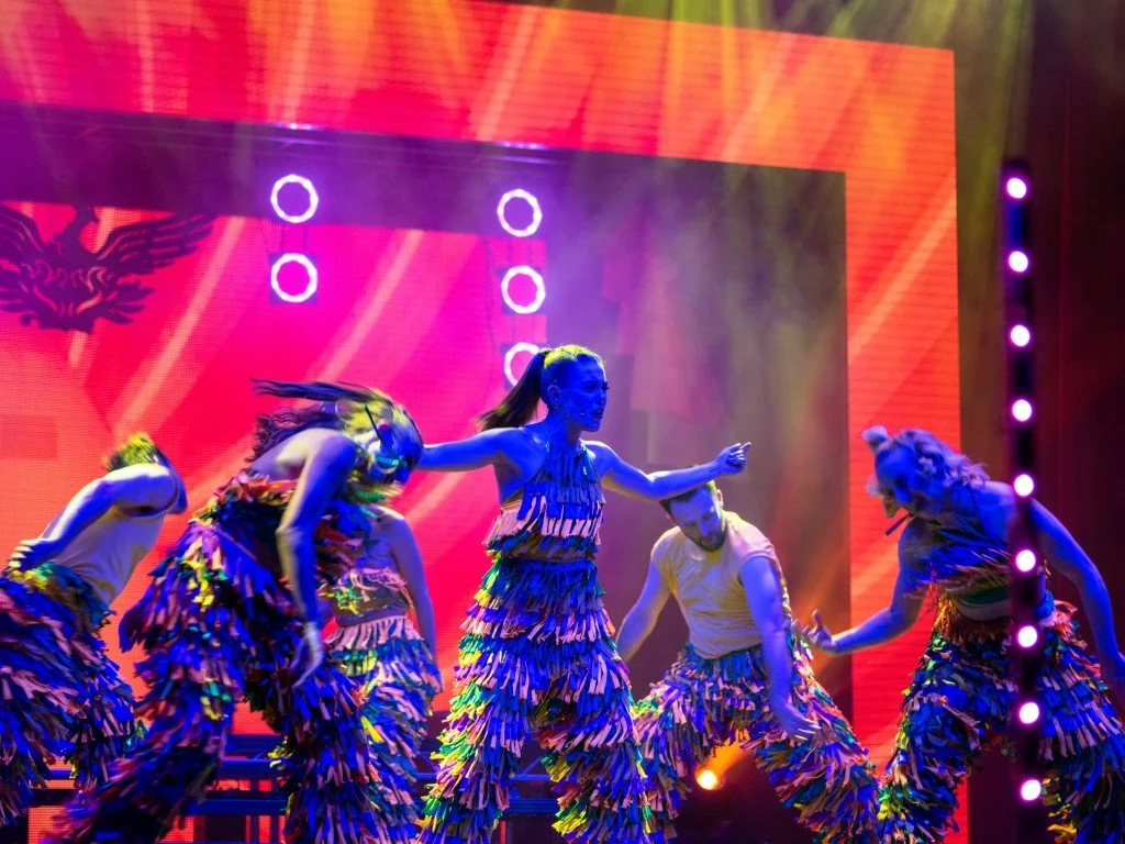 Group of dancers performing on stage, wearing colorful fringed costumes with neon lighting and a bright red backdrop.