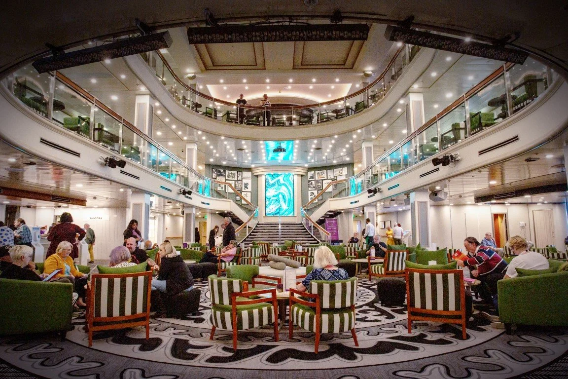 Interior view of a spacious, multi-level lounge with green and striped chairs, people sitting and chatting, a grand staircase, and decorative lighting and artwork.