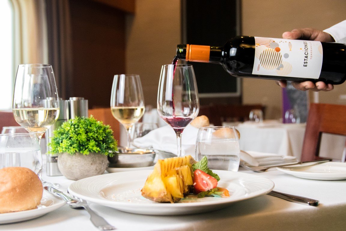 Wine being poured into a glass at a fine dining restaurant table with a plate of food, glasses of water, and a small green plant.