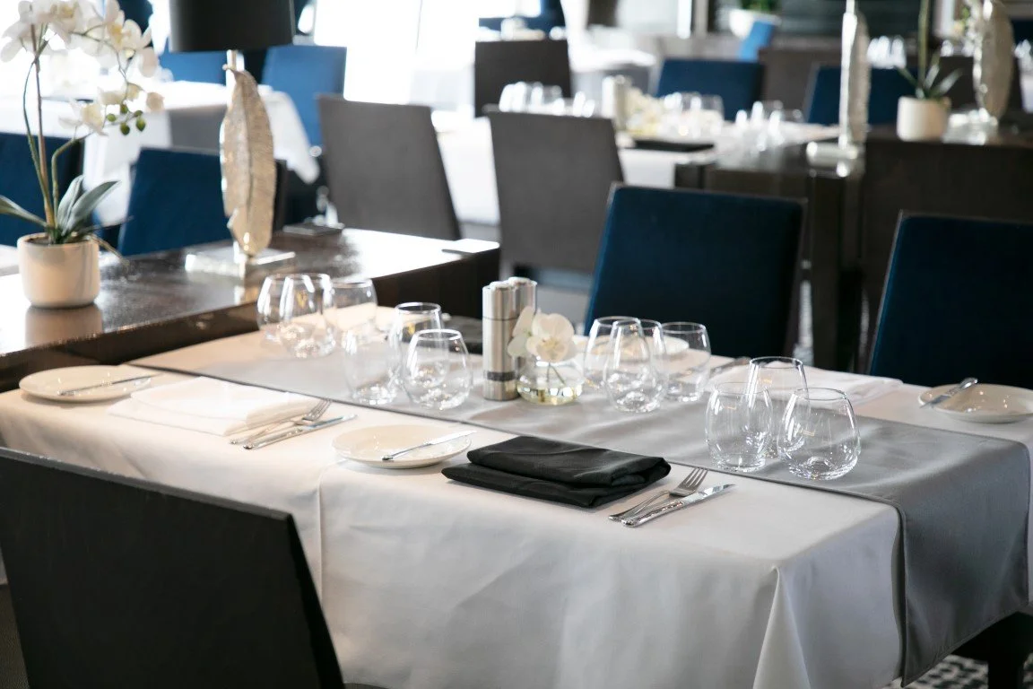 A formal dining table set with white tablecloth, black napkins, multiple wine glasses, water glasses, and silverware, decorated with a small white flower in a vase and a salt and pepper shaker, in a modern restaurant setting with additional tables and chairs in the background.