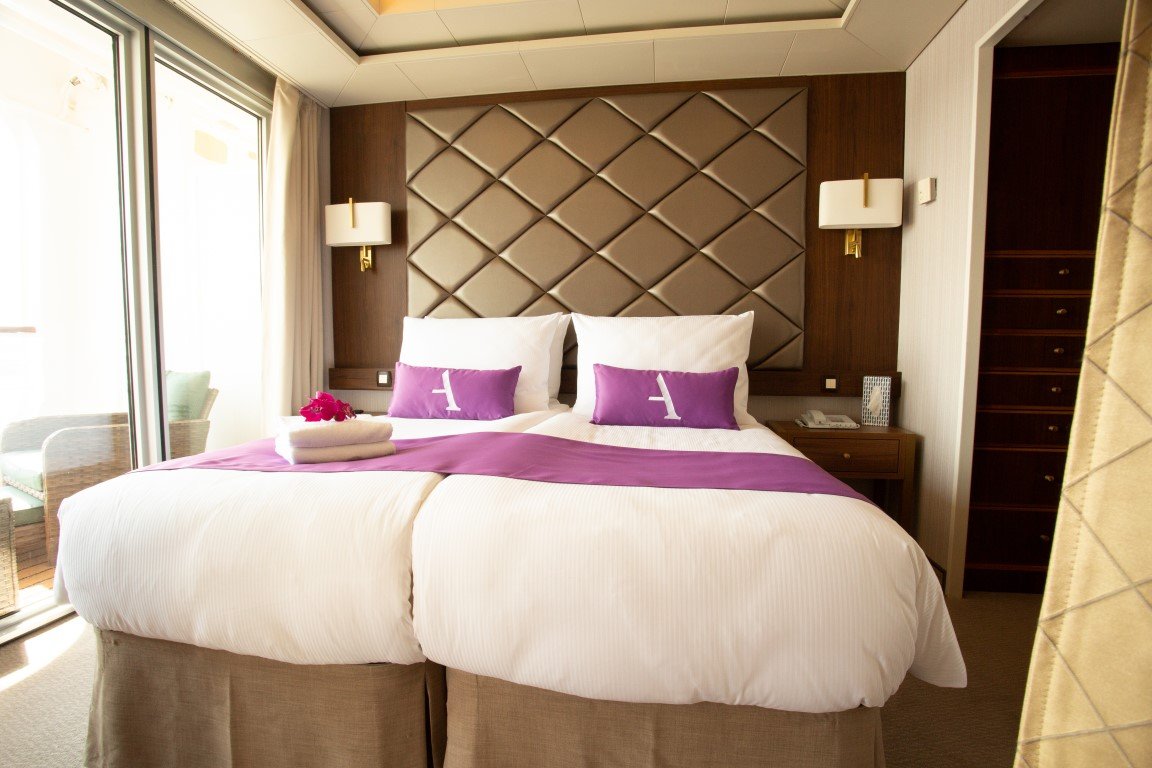A hotel room with a large bed featuring white linens, purple accent pillows with a white letter 'A', and a purple runner. There are two wall-mounted lamps on either side of the bed, and a towel with a flower on top is on the bed. The room has sliding glass doors leading to a balcony with outdoor seating, and a built-in shelving unit on the right.