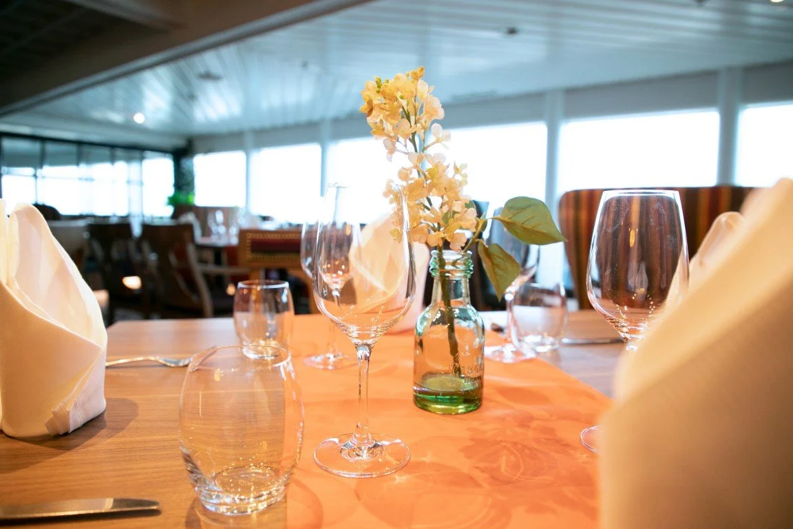 Elegant restaurant table setting with wine glasses, water glasses, folded napkins, and a small floral centerpiece on an orange table runner, with large windows in the background letting in natural light.