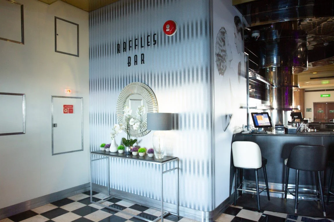 Interior of a bar named 'Rafaeles Bar' with black and white checkered floor, decorative mirror, vase with white orchids, and bar stools at the counter.