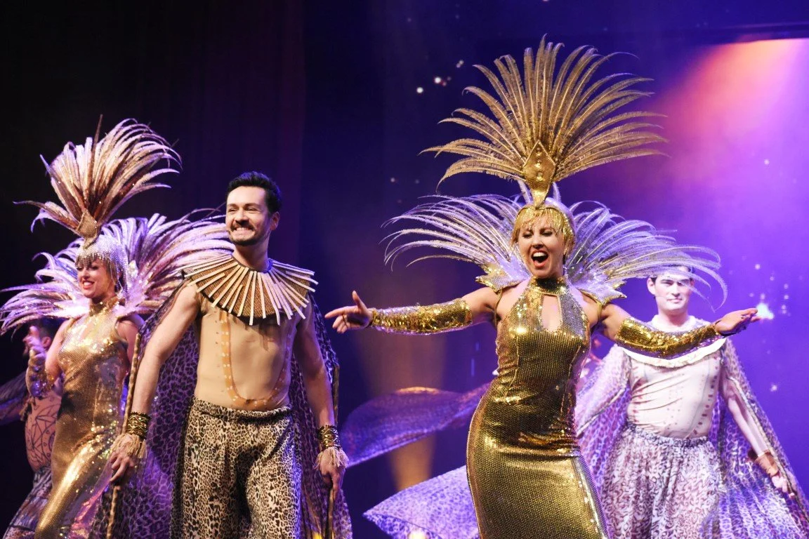 People dressed in shiny gold costumes with large feathered headdresses dancing on stage in a performance.