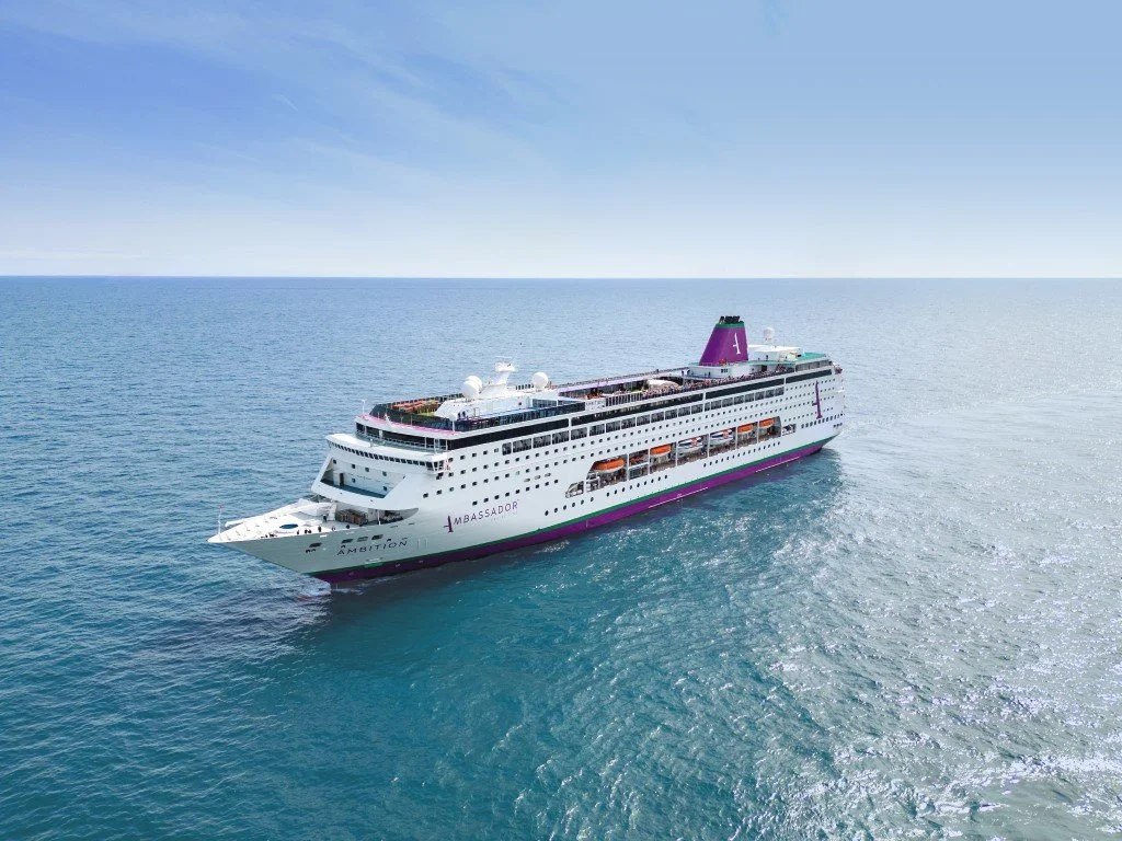 A large white cruise ship with purple and green accents sailing in the ocean under a partly cloudy sky.