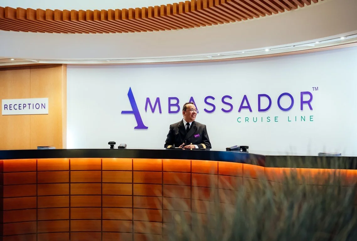 A man dressed in a cruise ship uniform standing at the reception desk of a cruise line, with a large sign that reads 'AMBASSADOR CRUISE LINE' behind him and a smaller sign that says 'RECEPTION' to the left.