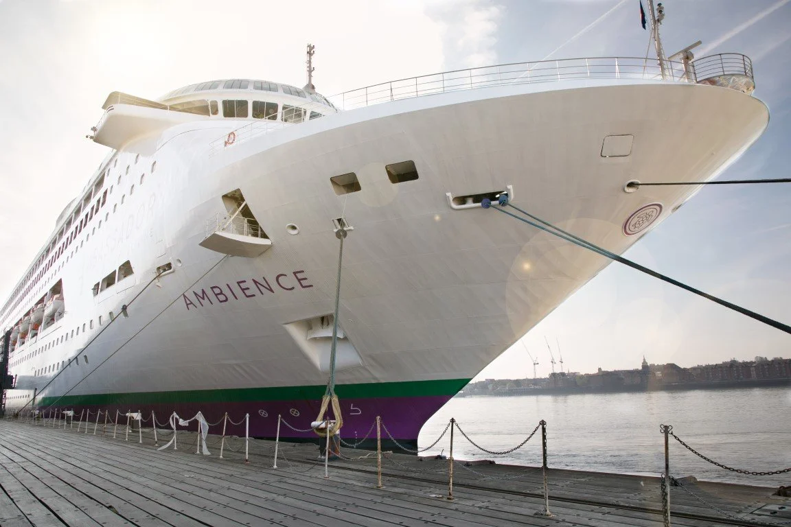 Large white cruise ship docked at a port with name 'AMBiance' painted on the side, moored with ropes, overlooking a river or ocean, with a city skyline and cranes in the distance