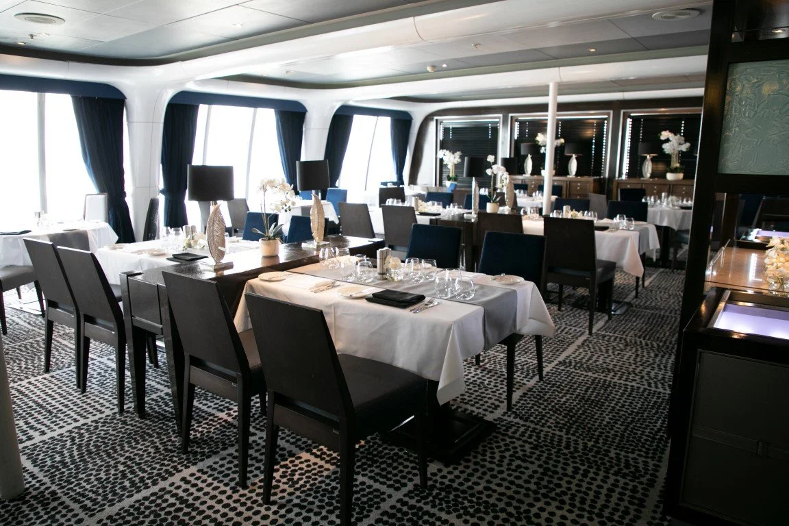Elegant restaurant with white tablecloth-covered tables, black chairs, and floral centerpieces, on a patterned black and white carpet with large windows and dark curtains.