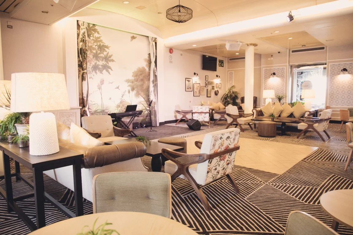 A spacious, well-lit lounge with seating, lamps, plants, and a small stage with musical instruments, decorated in neutral tones and modern style.