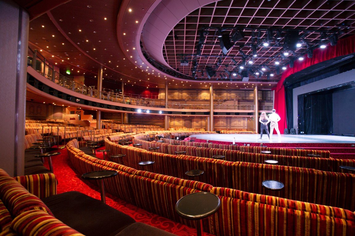An empty theater with red and striped upholstered seats, a stage with performers, and a balcony area above.
