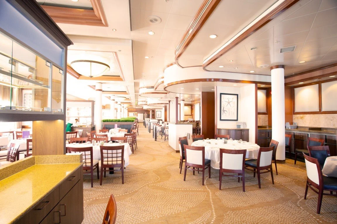 An elegantly decorated restaurant with round tables covered in white tablecloths and arranged with dining ware, high-backed chairs, a cozy and upscale ambiance with wooden accents, framed artwork, and a well-lit interior.