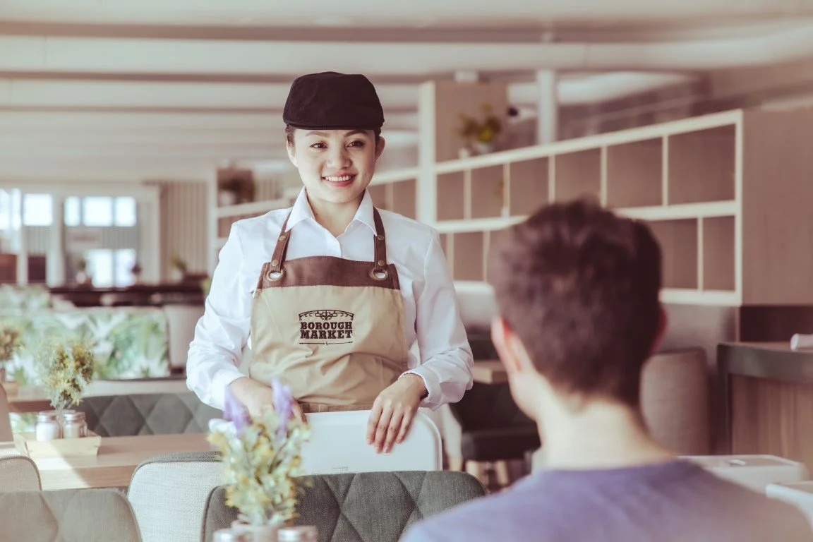 A smiling waitress wearing a beige apron and black hat takes an order from a male customer in a modern restaurant.
